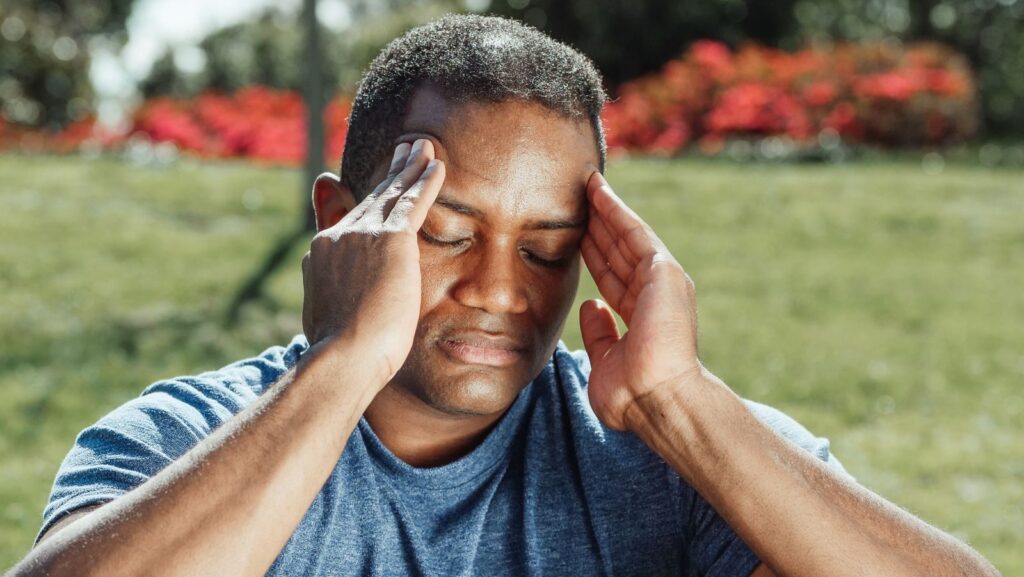 An adult man with eyes closed touches his temples, indicating a headache while seated outdoors.